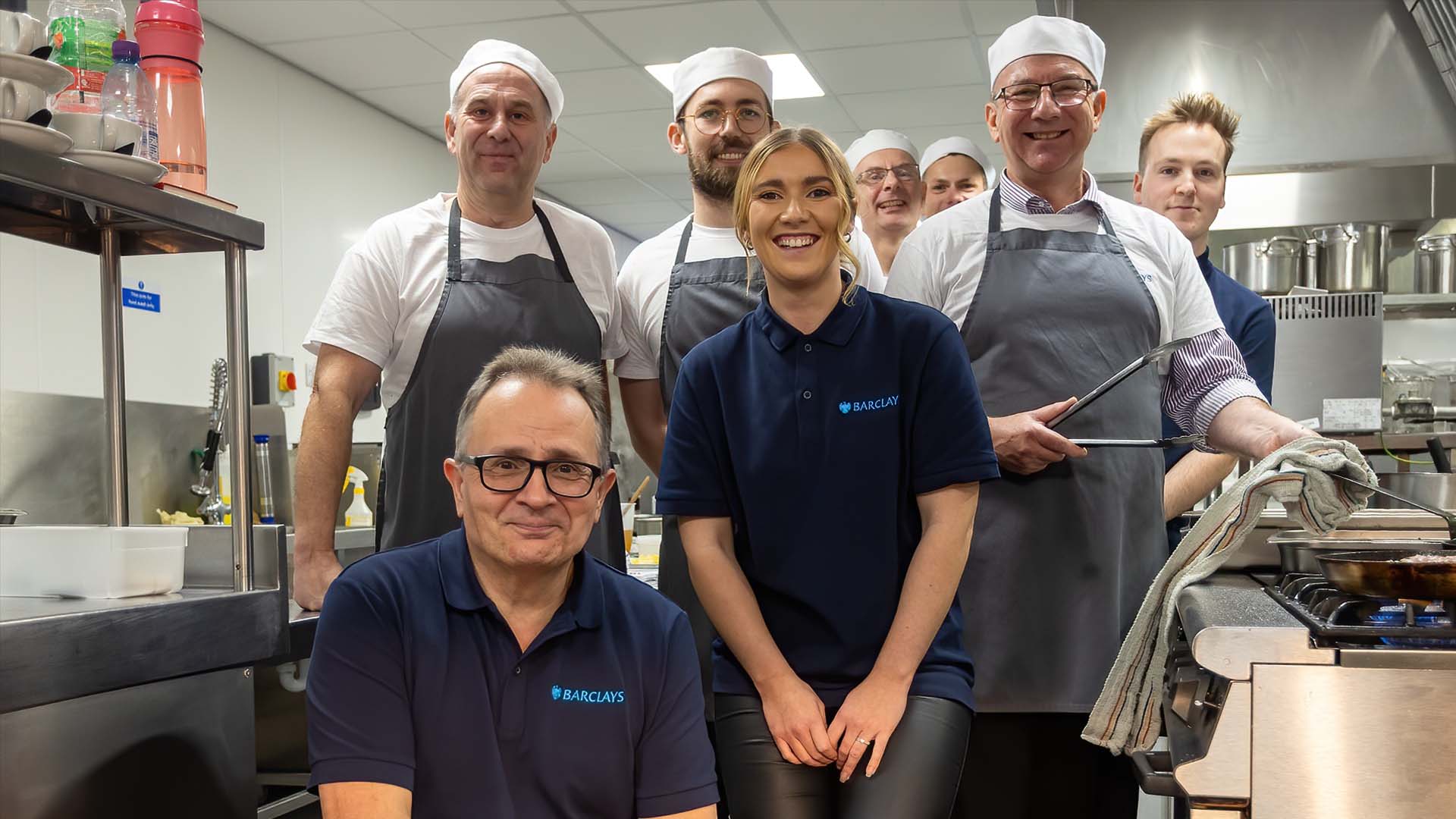 8 people in a kitchen, some wearing a chef's uniform, smiling at the camera